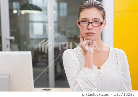 Woman wearing glasses and blouse touching chin sitting at desk with monitor glass wall, copy space Woman wearing glasses and blouse touching chin sitting at desk with monitor glass wall, copy space 135716282