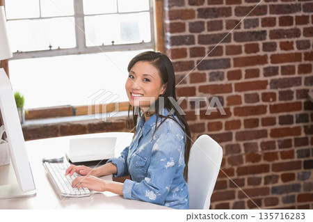 Asian woman sitting at loft desk typing on white keyboard with all-in-one monitor, denim shirt Asian woman sitting at loft desk typing on white keyboard with all-in-one monitor, denim shirt 135716283