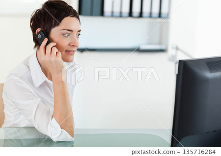 Mid-adult female in white shirt holding phone, looking at monitor at glass desk in office Mid-adult female in white shirt holding phone, looking at monitor at glass desk in office 135716284