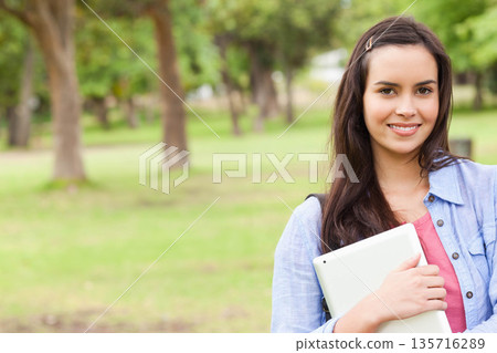 Teen girl standing holding tablet in park blue shirt coral top backpack-strap hair-clip, copy space 135716289