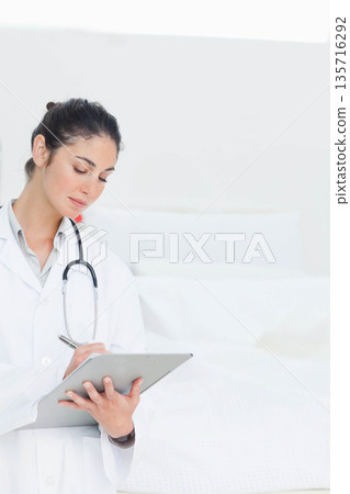 Female clinician in lab coat writing notes on clipboard at exam bed with stethoscope, copy space 135716292