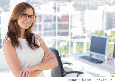Mid-adult woman posing and smiling by windows, wearing glasses, desk monitor visible, copy space Mid-adult woman posing and smiling by windows, wearing glasses, desk monitor visible, copy space 135716296