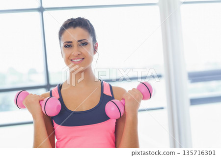Female holding pink dumbbells near shoulders in workout studio with grid windows, wearing pink tank 135716301