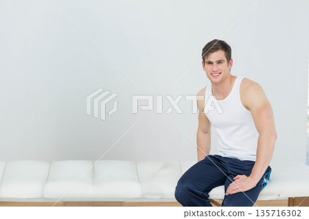 Man sitting on white tufted bench by blinds in studio, wearing tank and pants, copy space 135716302
