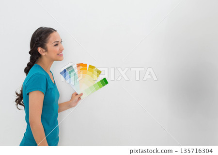 Woman in 20s standing left of home wall holding, inspecting paint swatches, teal shirt, copy space Woman in 20s standing left of home wall holding, inspecting paint swatches, teal shirt, copy space 135716304