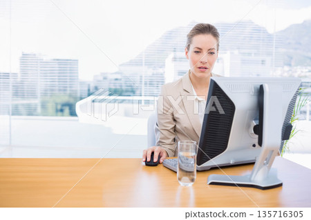Mid adult woman in beige blazer working at office desk with monitor, mouse, glass of water Mid adult woman in beige blazer working at office desk with monitor, mouse, glass of water 135716305