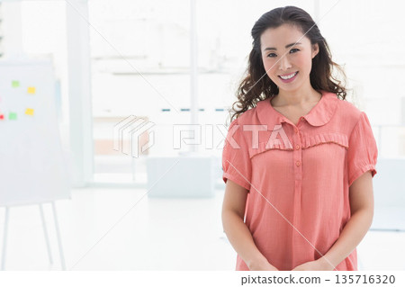 Asian woman standing and smiling in bright studio wearing coral blouse near whiteboard, copy space Asian woman standing and smiling in bright studio wearing coral blouse near whiteboard, copy space 135716320