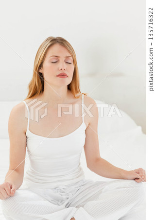 Woman meditating crosslegged on white bedding in pale bedroom wearing camisole and pajama pants Woman meditating crosslegged on white bedding in pale bedroom wearing camisole and pajama pants 135716322
