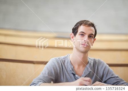 Adult male student sitting in lecture hall with wooden desks, taking notes with pen, wearing henley Adult male student sitting in lecture hall with wooden desks, taking notes with pen, wearing henley 135716324