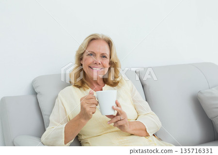 Senior female sitting on light-gray sofa at home holding white ceramic mug with both hands Senior female sitting on light-gray sofa at home holding white ceramic mug with both hands 135716331