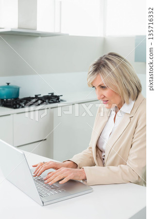 Woman in beige blazer typing on silver laptop at kitchen counter with teal pot, copy space Woman in beige blazer typing on silver laptop at kitchen counter with teal pot, copy space 135716332