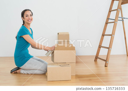 20s woman kneeling on wood floor packing moving boxes with tape dispenser, ladder, copy space 135716333