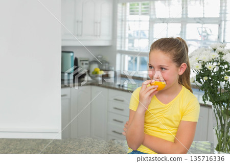 Female child sitting on kitchen counter wearing yellow tee holding half orange biting, copy space 135716336