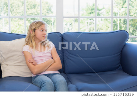 Female child sitting with crossed arms on deep-blue sofa by multi-pane window showing greenery Female child sitting with crossed arms on deep-blue sofa by multi-pane window showing greenery 135716339
