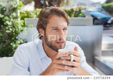 Man in 30s wearing light blue shirt, holding white mug while sitting at patio with planters Man in 30s wearing light blue shirt, holding white mug while sitting at patio with planters 135716344