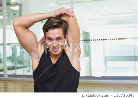 Man stretching overhead arm in fitness studio wearing black tank top by mirror and glass partition Man stretching overhead arm in fitness studio wearing black tank top by mirror and glass partition 135716345