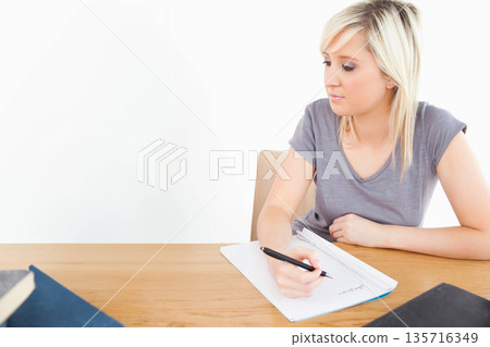 Female student writing in lined notebook at table holding black pen, hardcovers nearby, copy space Female student writing in lined notebook at table holding black pen, hardcovers nearby, copy space 135716349