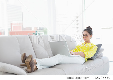 Adult Asian woman reclining on sofa in living room wearing yellow blouse using laptop, copy space Adult Asian woman reclining on sofa in living room wearing yellow blouse using laptop, copy space 135716350