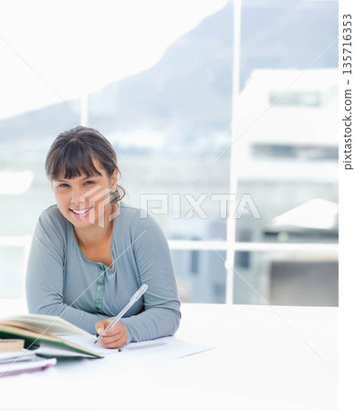 Woman leaning forward, holding pen over notebook and binder, studying at white table near windows Woman leaning forward, holding pen over notebook and binder, studying at white table near windows 135716353