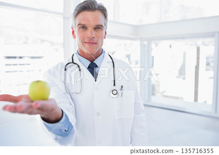 Male doctor standing in clinic offering green apple wearing lab coat and stethoscope Male doctor standing in clinic offering green apple wearing lab coat and stethoscope 135716355