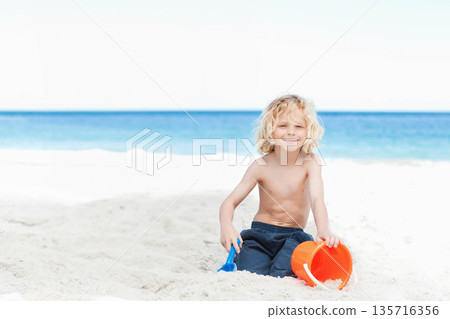 Child boy sitting shirtless on white sand by ocean, playing with orange bucket and blue shovel 135716356