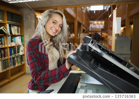 Adult woman in plaid shirt lifting copier lid and placing spiral notebook at library near shelves 135716363