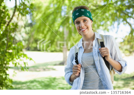 Woman standing in park holding backpack straps, wearing green bandana and grey tank, copy space 135716364