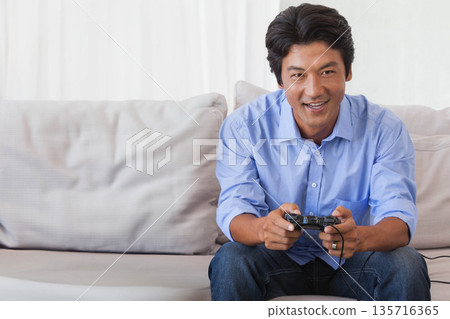Mid adult Asian man wearing light blue shirt and dark jeans, playing wired controller on sofa Mid adult Asian man wearing light blue shirt and dark jeans, playing wired controller on sofa 135716365