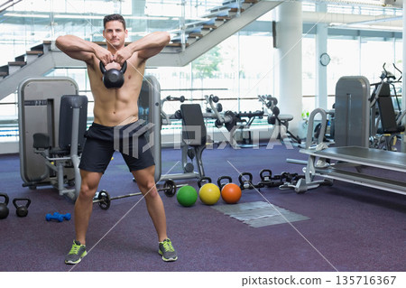 Man standing shirtless holding black kettlebell at chest height in modern gym wearing black shorts 135716367