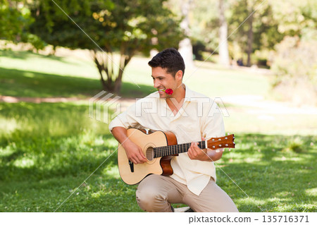 Mid-adult man sitting on small stool in park playing acoustic guitar holding red rose in mouth Mid-adult man sitting on small stool in park playing acoustic guitar holding red rose in mouth 135716371