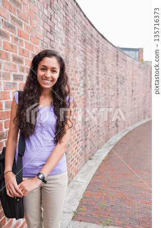 Woman posing beside curved red-brick wall on pathway, wearing lilac top, with black bag, copy space Woman posing beside curved red-brick wall on pathway, wearing lilac top, with black bag, copy space 135716373
