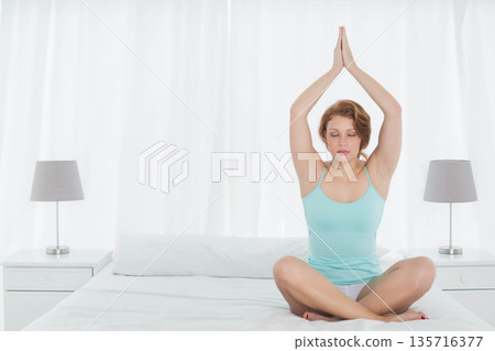 Female meditating in turquoise tank top and white shorts on bed with white bedding, copy space 135716377