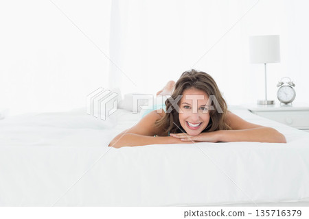 Adult woman propped on arms smiling at camera on white bed wearing blue tank, nightstand clock 135716379