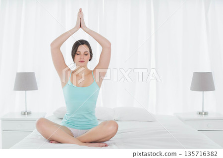 Adult female sitting crosslegged on bed meditating with hands overhead, wearing light blue tank top Adult female sitting crosslegged on bed meditating with hands overhead, wearing light blue tank top 135716382