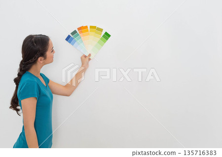 Single female teal tee on left, holding, comparing paint swatches on plain white wall, copy space Single female teal tee on left, holding, comparing paint swatches on plain white wall, copy space 135716383