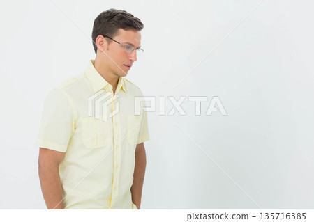 Man wearing pale yellow shirt, clear glasses standing left on light wall, looking down, copy space Man wearing pale yellow shirt, clear glasses standing left on light wall, looking down, copy space 135716385