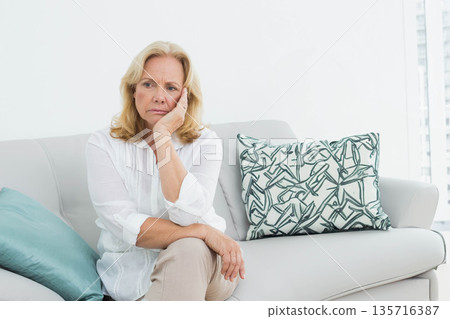 Senior woman sitting on gray sofa at home in white blouse, beige trousers beside aqua cushion 135716387