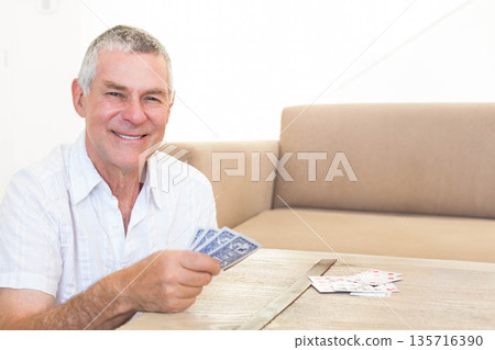 Senior man sitting at wooden table holding playing cards with beige sofa behind, copy space 135716390