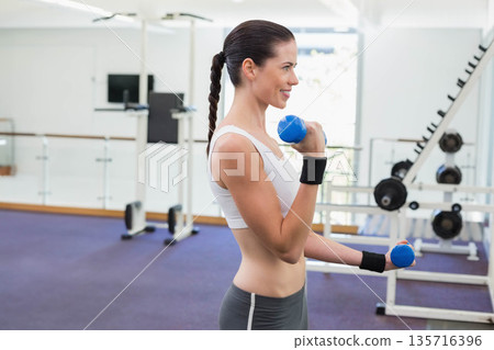 Female exerciser performing dumbbell curls in gym wearing white sports bra and gray leggings 135716396