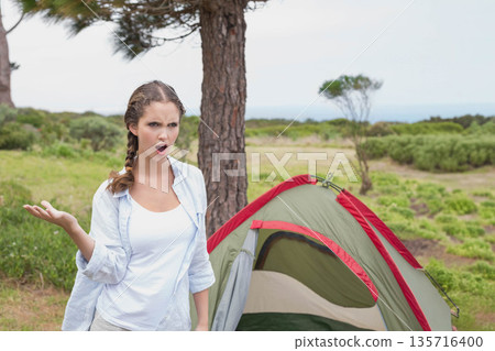 Woman wearing white tank and light shirt, standing gesturing by tent and pine trunk at coast 135716400