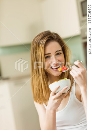 Woman in twenties wearing white top, holding white bowl and lifting spoon at kitchen counter Woman in twenties wearing white top, holding white bowl and lifting spoon at kitchen counter 135716402