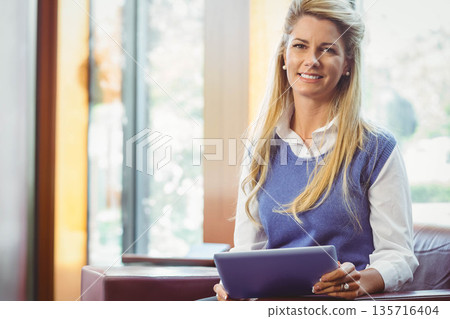 Mid-adult female sitting on leather chair holding tablet in bright office in shirt and sweater vest 135716404