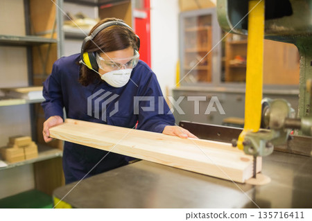 Female woodworker wearing navy coveralls and safety gear guiding long plank at band saw in workshop Female woodworker wearing navy coveralls and safety gear guiding long plank at band saw in workshop 135716411