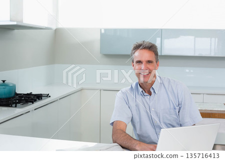 Middle-aged man in blue shirt sitting at kitchen counter using laptop beside teal pot, copy space Middle-aged man in blue shirt sitting at kitchen counter using laptop beside teal pot, copy space 135716413