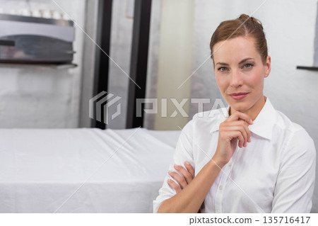 Mid-adult woman standing hand under chin in white shirt in treatment room near table, copy space 135716417