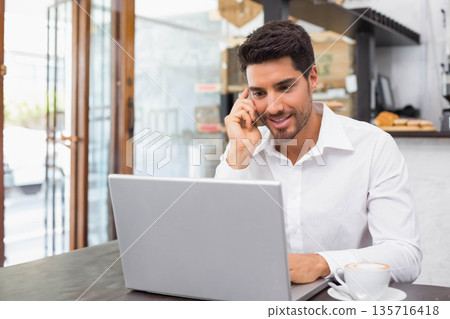 Adult male wearing shirt sitting at wooden table cafe using laptop, speaking on phone with latte Adult male wearing shirt sitting at wooden table cafe using laptop, speaking on phone with latte 135716418