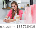 Female sitting at cafe table wearing pink cardigan, holding smartphone with latte and shopping bags 135716419
