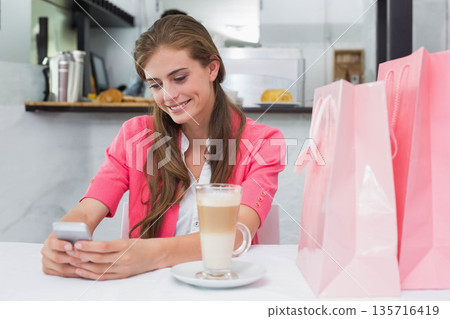 Female sitting at cafe table wearing pink cardigan, holding smartphone with latte and shopping bags 135716419
