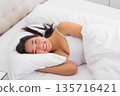 Asian woman lying on right-side in bed, smiling, resting head on pillow, in camisole under duvet 135716421