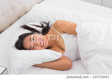 Asian woman lying on right-side in bed, smiling, resting head on pillow, in camisole under duvet Asian woman lying on right-side in bed, smiling, resting head on pillow, in camisole under duvet 135716421